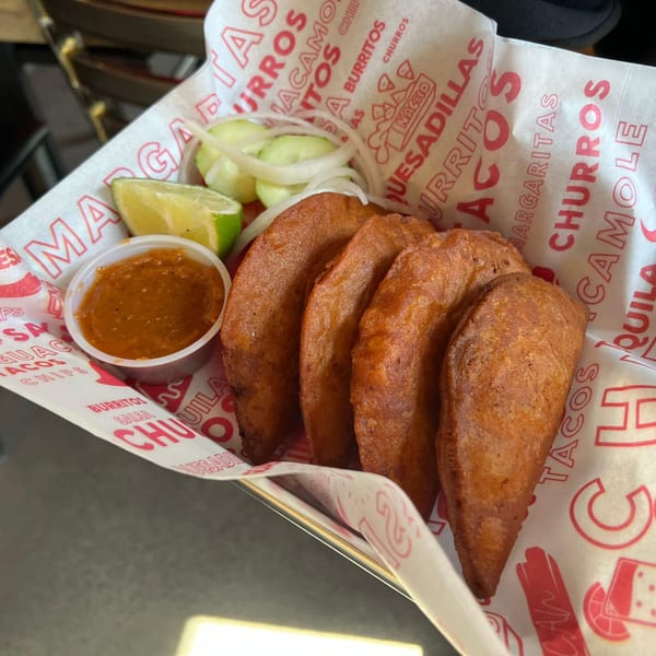 Four crispy tacos with lime, cucumber, onion, and salsa in a basket lined with branded paper.