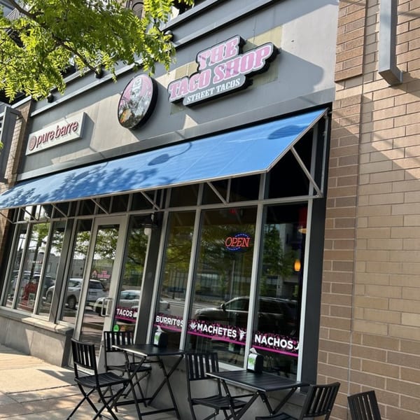 The Taco Shop storefront with a blue awning, outdoor seating, and menu items displayed on windows.