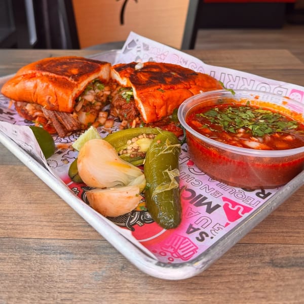 A birria torta with a side of consommé, pickled onions, jalapeños, and lime on a tray.