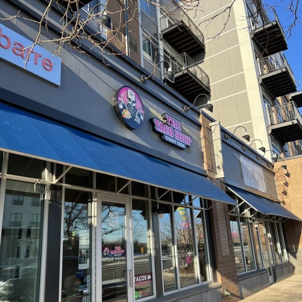 The Taco Shop storefront with blue awnings, large windows, and a skull logo sign.